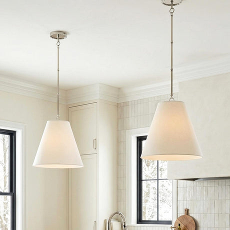 A transitional kitchen features two Luca Medium 15" Conical Pendants, Polished Nickel with white linen shades along the ceiling over a cream backsplash and cabinetry.