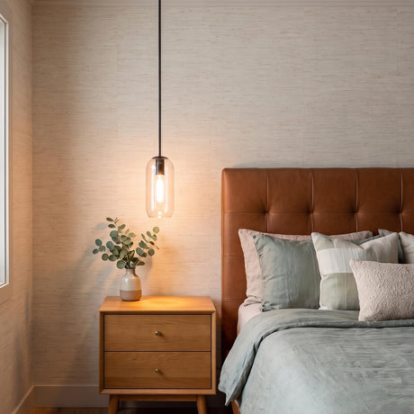 A mid-century bedroom with a matte black pendant and clear capsule glass hanging over a light wood nightstand beside a tufted brown leather headboard.