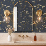 A transitional bathroom with two satin brass and clear glass wall sconces flanking an arched gold mirror against navy botanical wallpaper and travertine counters.