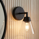 A modern bathroom with an Owen 1 Light Vanity Wall Sconce, Matte Black featuring a tapered glass shade mounted on fluted cream tiles beside a round mirror.