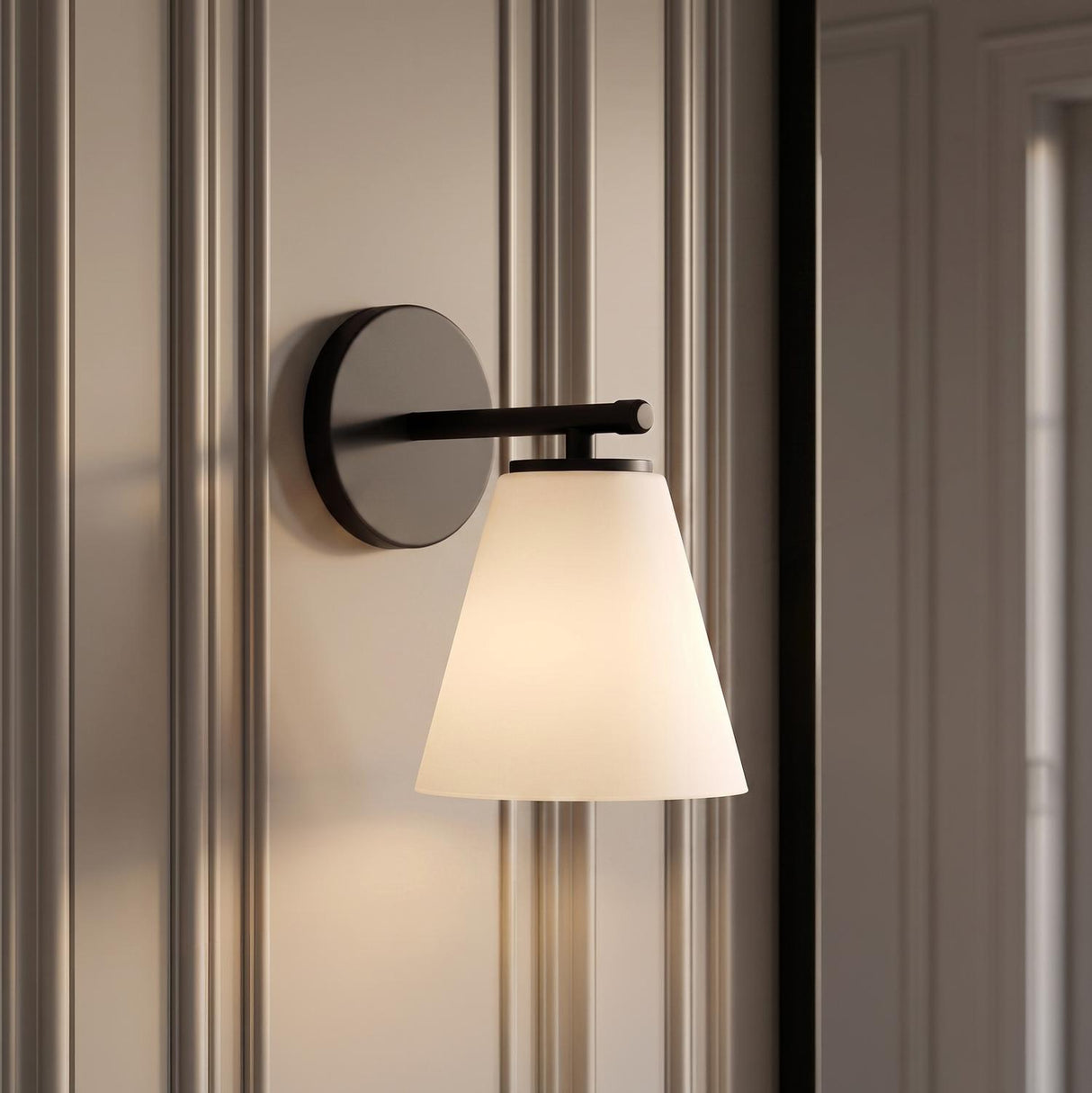 A transitional bathroom featuring a Carlisle Vanity Wall Sconce, Matte Black with Opal Glass mounted on a neutral paneled wall with decorative vertical molding details.