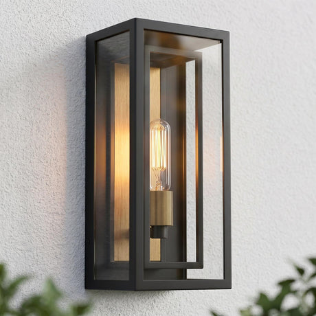 A modern home exterior featuring the Jaxon Outdoor Wall Light, a matte black and brass wall lantern glowing at dusk on a white stucco wall with green foliage.
