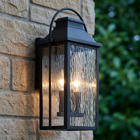 A traditional stone wall features the Preston Outdoor Wall Light, Medium, in a textured black finish glowing at dusk against a backdrop of green landscaping.