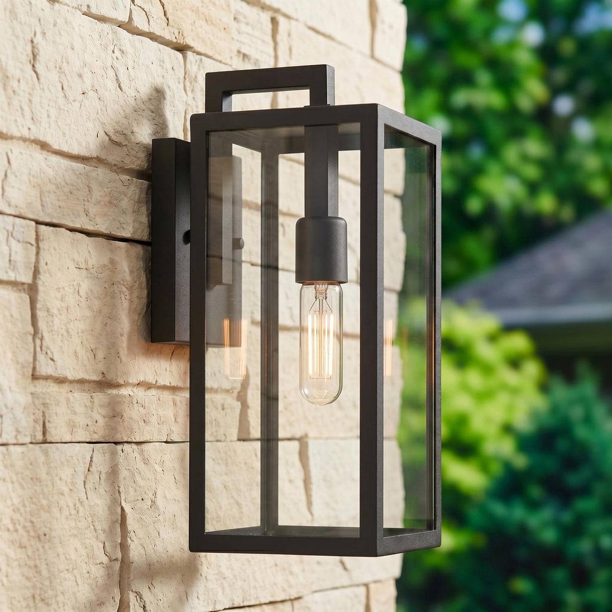 A modern home exterior with a matte black Sam Outdoor Wall Light, Small 15" lantern glowing on a stacked stone wall beside green garden landscaping.