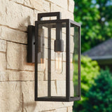A modern home exterior with a matte black Sam Outdoor Wall Light, Small 15" lantern glowing on a stacked stone wall beside green garden landscaping.