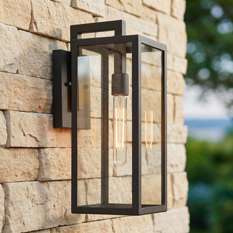 A modern home exterior with a textured black Sam Outdoor Wall Light, Medium 18" glowing on a tan stacked stone wall beside blurred green landscaping at twilight.