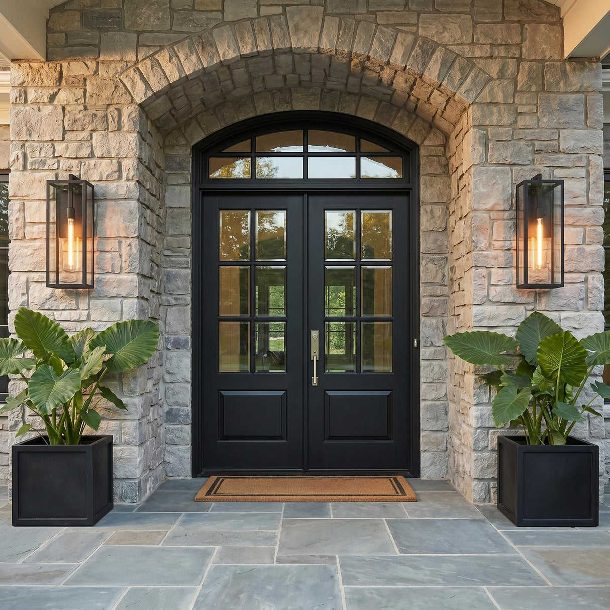 A transitional stone home entry with two Sam Outdoor Wall Light, Large 24" in matte black flanking a black double door with large leafy plants at dusk.