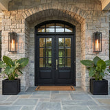 A transitional stone home entry with two Sam Outdoor Wall Light, Large 24" in matte black flanking a black double door with large leafy plants at dusk.