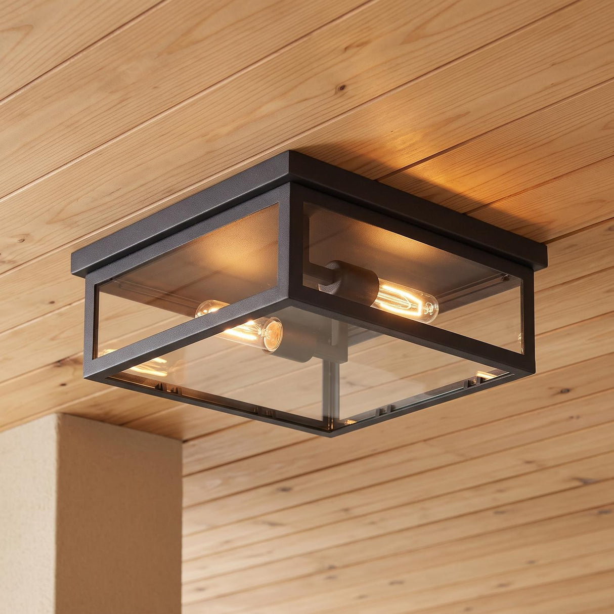 A modern porch featuring a Sam Outdoor Flush Mount in matte black finish glowing on a light wood plank ceiling beside a stucco pillar at dusk.