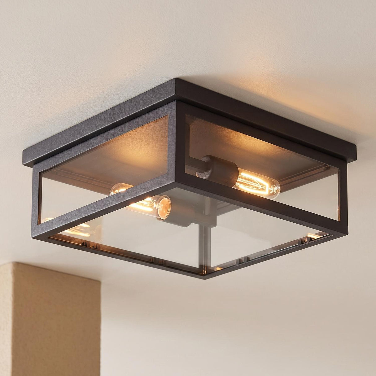 A modern farmhouse porch with a matte black Sam Outdoor Flush Mount lantern glowing on a white ceiling next to a beige stucco pillar at night.