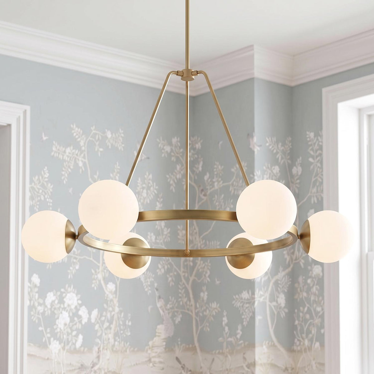 A transitional dining room featuring the Castell 6 Globe Round Chandelier 36", Aged Brass suspended before soft blue botanical wallpaper and white crown molding.