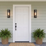 A modern farmhouse entry featuring light green siding and two Glenn Outdoor Wall Light, Matte Black fixtures glowing while flanking a white front door above potted ferns.