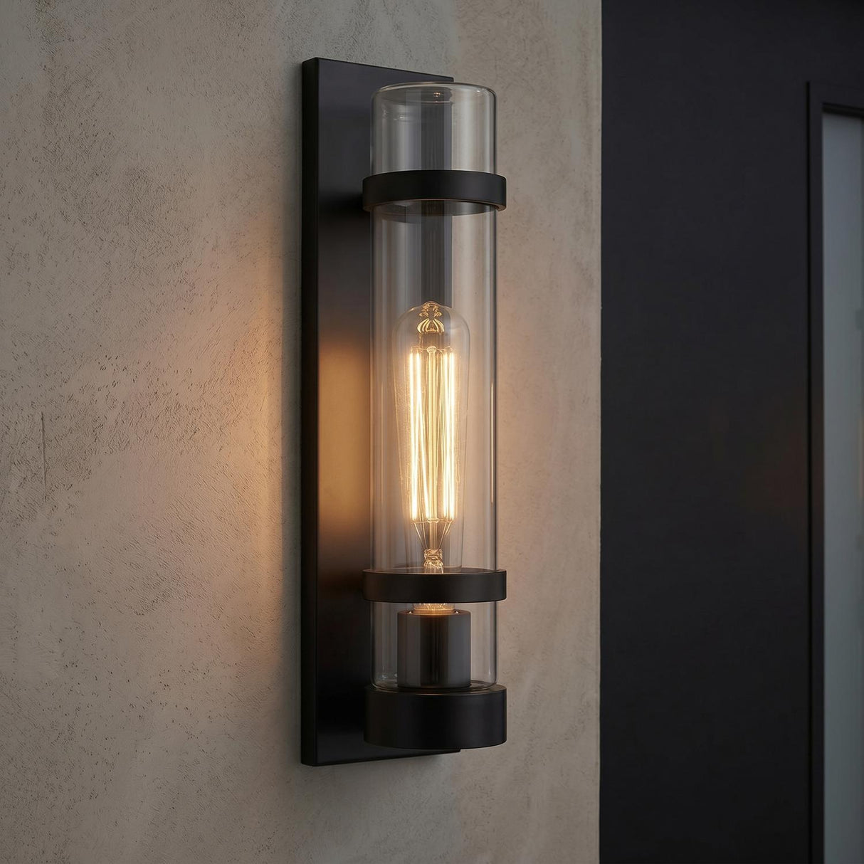 A modern outdoor entryway featuring the Glenn Outdoor Wall Light, Matte Black with a clear glass cylinder and filament bulb mounted on a textured beige wall.