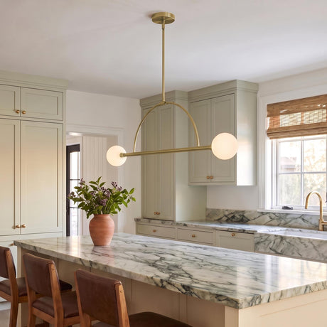 A transitional kitchen featuring the Milo 2 Globe Linear Arc Pendant in aged brass with white globes over a marble island and sage green cabinetry.