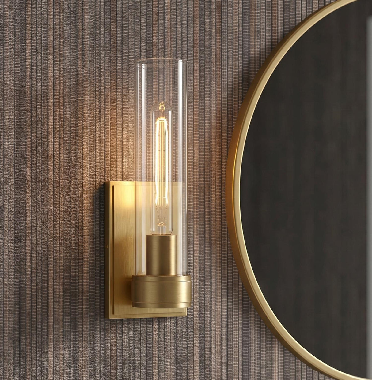 A contemporary bathroom featuring a Sutton 1 Light Wall Sconce, Satin Brass with a clear glass cylinder flanking a round gold-rimmed mirror on textured wallpaper.