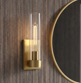 A contemporary bathroom featuring a Sutton 1 Light Wall Sconce, Satin Brass with a clear glass cylinder flanking a round gold-rimmed mirror on textured wallpaper.