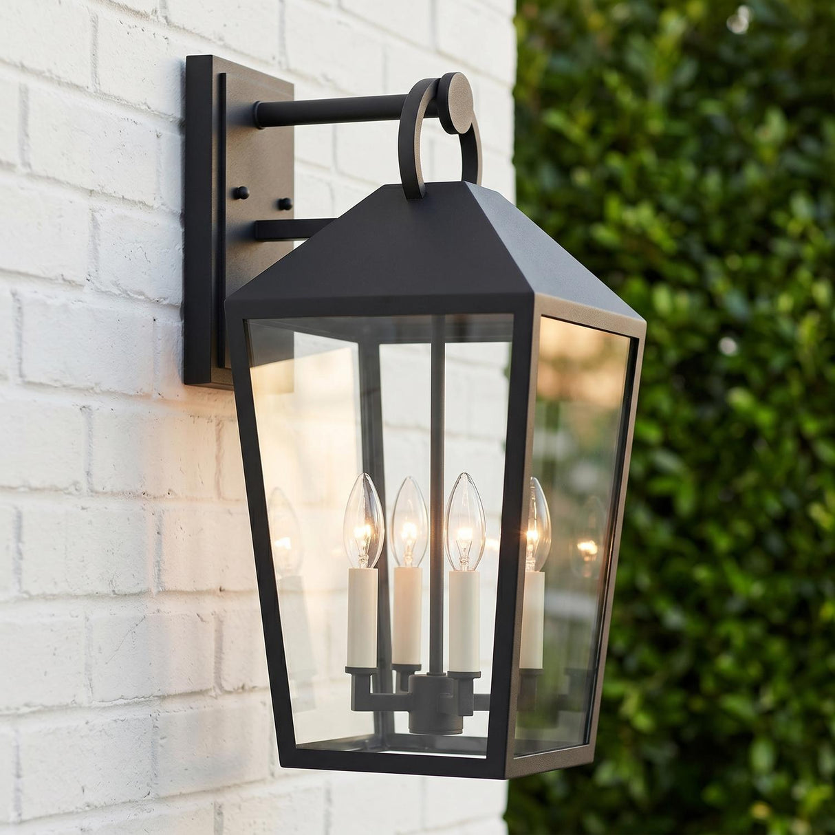 A modern farmhouse exterior with the matte black Cole Outdoor Wall Light, Large 21.5" Black, glowing at dusk on a white brick wall near green hedges.
