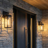 A contemporary outdoor entryway featuring two matte black and clear glass Cyrus Outdoor Wall Light, Medium, sconces flanking a dark wood door on a gray stone wall.