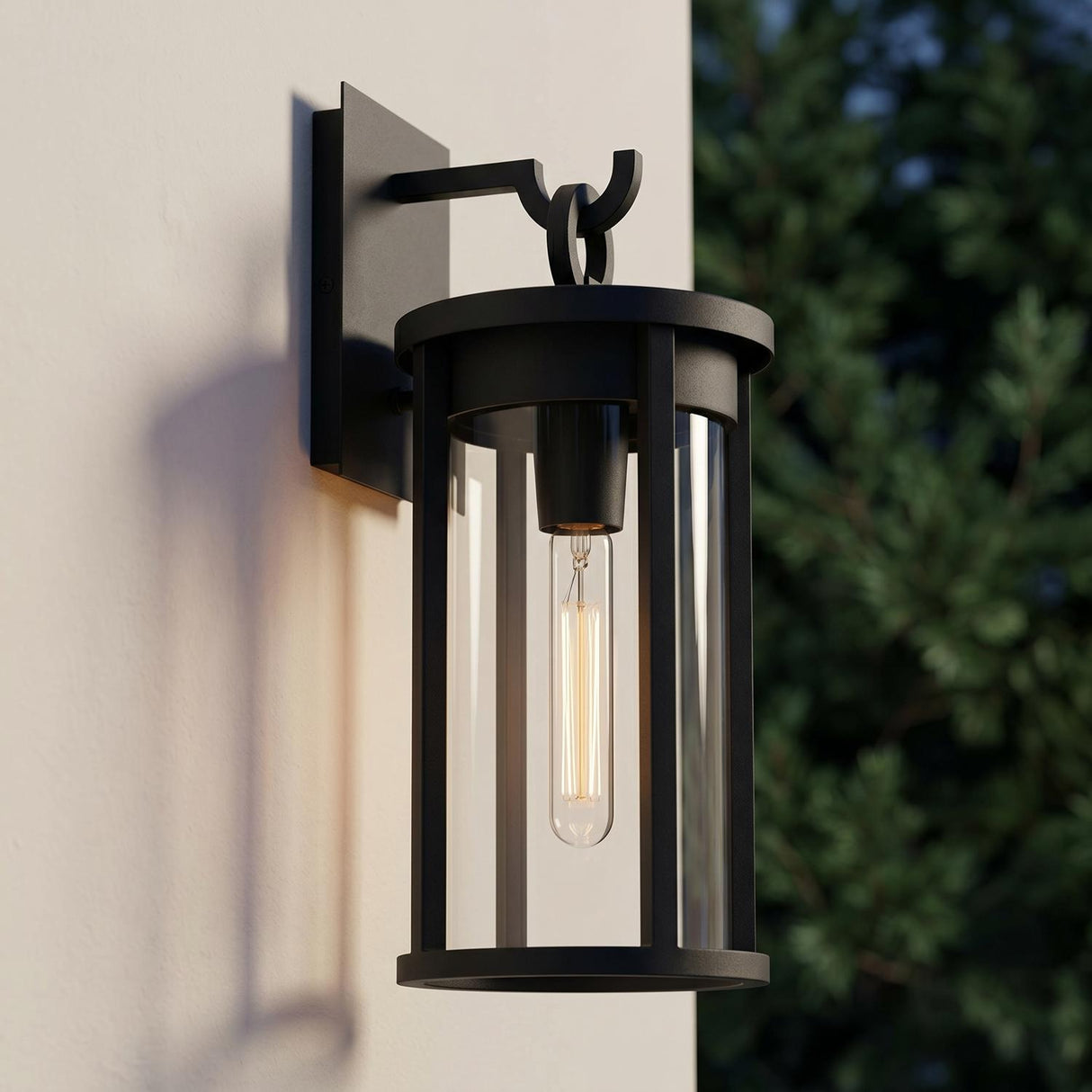 A modern home exterior featuring the matte black Cyrus Outdoor Wall Light, Medium mounted on a white stucco wall at twilight with dark evergreen trees in the background.