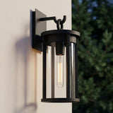 A modern home exterior featuring the matte black Cyrus Outdoor Wall Light, Medium mounted on a white stucco wall at twilight with dark evergreen trees in the background.
