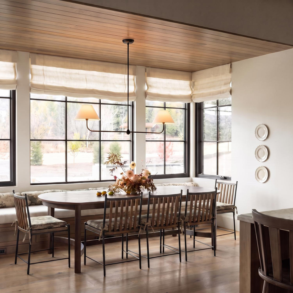 A transitional dining room featuring the matte black Holden 2 Shade Linear Pendant over a wooden table under a planked ceiling and windows with cream roman shades.