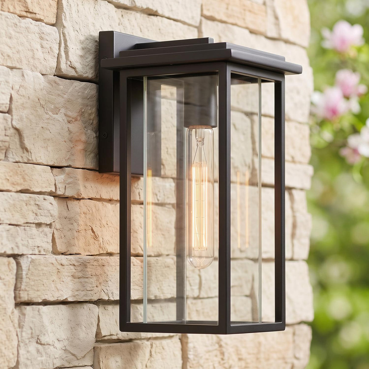 A modern home exterior featuring the Colin Outdoor Wall Light, Small in matte black glowing on a tan stacked stone wall beside flowering pink landscaping.