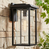 A modern home exterior featuring a matte black Colin Outdoor Wall Light, Medium wall lantern glowing on a tan stone wall at dusk beside green foliage.