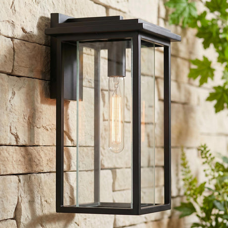 A modern home exterior featuring a matte black Colin Outdoor Wall Light, Medium wall lantern glowing on a tan stone wall at dusk beside green foliage.