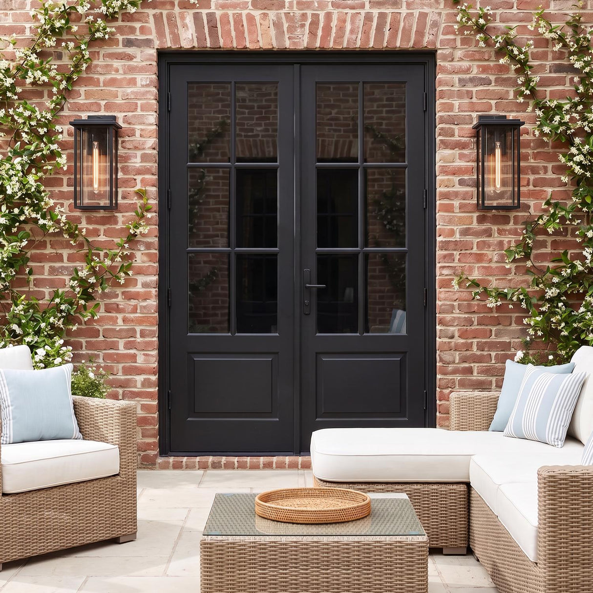 A traditional red brick patio features two matte black Colin Outdoor Wall Light Large lanterns flanking black French doors, accented by climbing jasmine and wicker furniture.