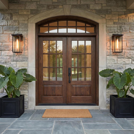 A transitional home entry with two matte black wall lanterns flanking dark wood double doors on a stone facade at twilight with large potted elephant ear plants.