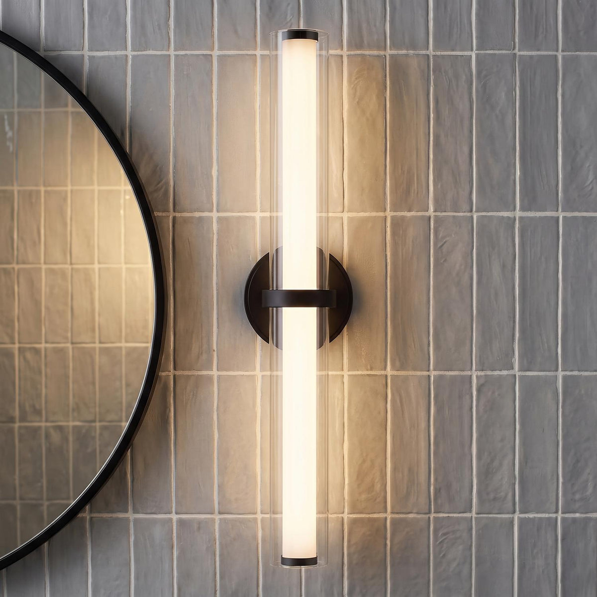A modern bathroom with a matte black Ellie LED Vanity Light featuring a clear glass cylinder mounted on gray vertical tiles beside a circular mirror.