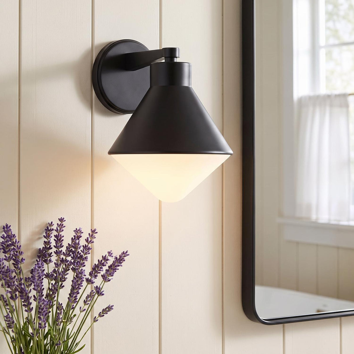 A modern farmhouse bathroom featuring an Andie Wall Sconce, Matte Black, with a frosted glass shade mounted on cream shiplap walls beside a black-framed mirror.