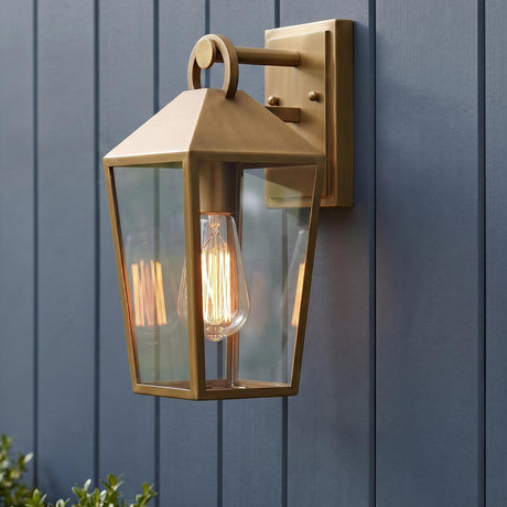 A modern farmhouse exterior with the Cole Outdoor Wall Light in burnished brass glowing on dark blue vertical siding at dusk near green boxwood landscaping.