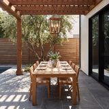 A Mediterranean patio with the Cole Outdoor Hanging Light in burnished brass suspended from a wood pergola over a dining set, featuring white stucco and an olive tree.