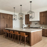 A modern kitchen features three Noa 4 Light Pendant 13", Matte Black with White lanterns over a marble island with walnut cabinets and leather stools.