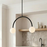 A minimalist kitchen featuring a matte black Risa 2 Globe Arc Pendant 26" with frosted glass globes over the sink area, textured backsplash, and natural wood shelf.