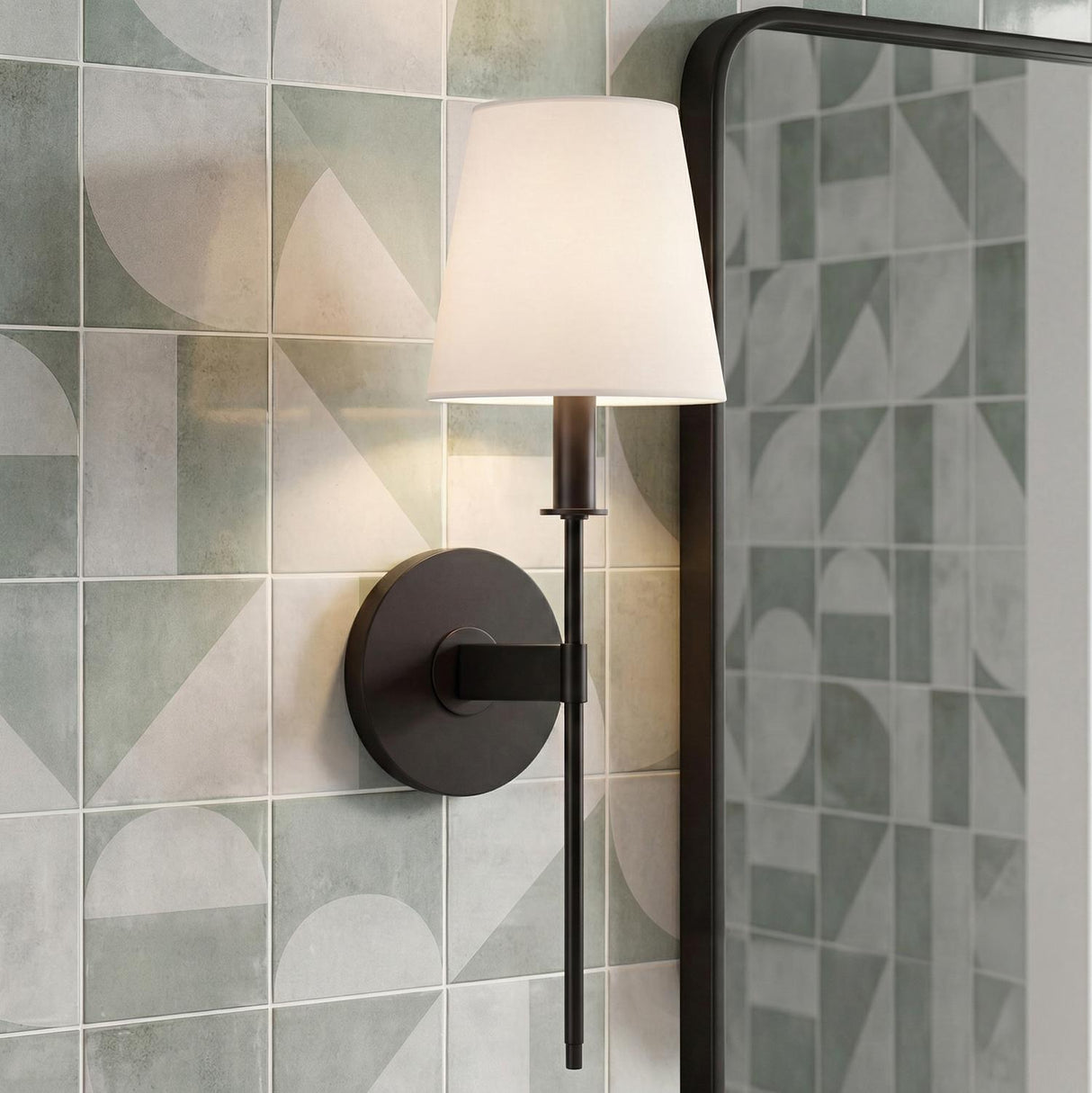 A contemporary bathroom features a Taylor Wall Sconce, Matte Black with a white linen shade installed on a geometric sage green tiled wall beside a mirror.