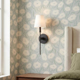 A transitional bedroom with the Taylor Wall Sconce in Matte Black featuring a white linen shade mounted on floral wallpaper beside a cream upholstered wingback headboard.