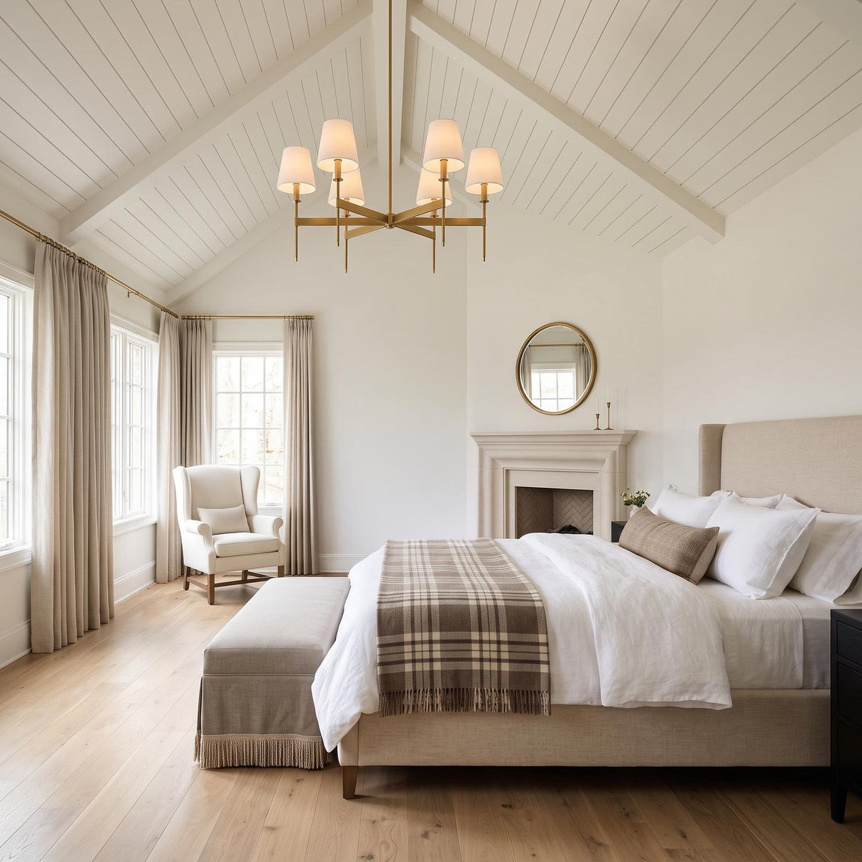 A modern farmhouse bedroom with the Taylor 6 Light Chandelier in aged brass with linen shades suspended from a vaulted beamed ceiling above a beige bed.