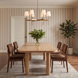 A transitional dining room featuring the Taylor 6 Light Chandelier 30" in Aged Brass with white linen shades over a light oak table and striped wallpaper.