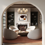 Taylor six light Chandelier in aged brass with white fabric shades, shown in a loving room with four rounded barrel chairs, modern art, and a marble fireplace.