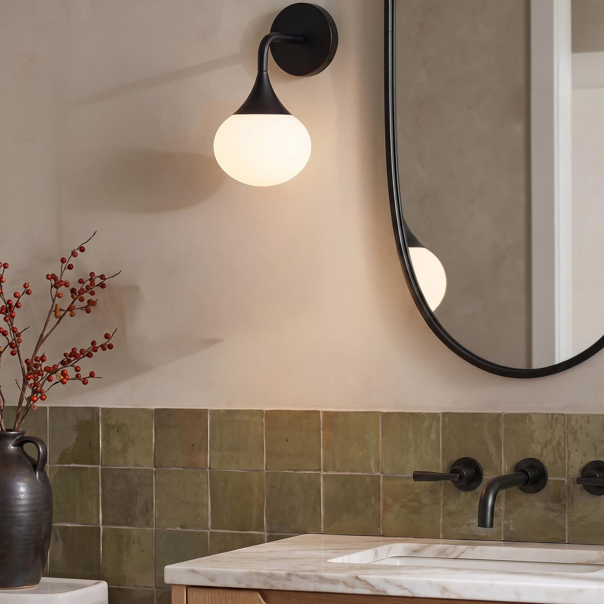A contemporary bathroom with a matte black Talia wall sconce and frosted glass globe mounted beside an oval mirror over a white marble vanity and green square tiles.