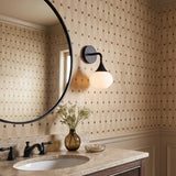 A transitional bathroom with a matte black and white glass Talia Wall Sconce mounted beside a round mirror above a tan marble vanity countertop.