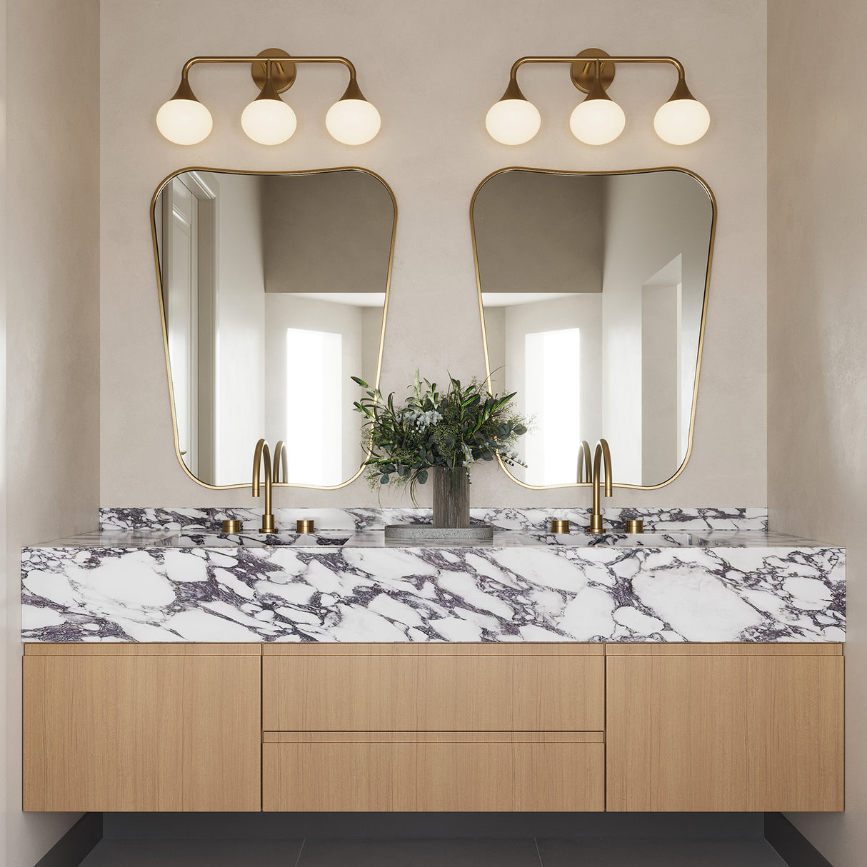 Two Talia 3 Light 24" LED Vanity lights, shown in aged brass finish with white opal glass globes, are in a primary bath with beige plaster walls, hung above two modern curved mirrors.