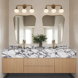 Two Talia 3 Light 24" LED Vanity lights, shown in aged brass finish with white opal glass globes, are in a primary bath with beige plaster walls, hung above two modern curved mirrors.