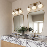 Two Talia 3 Light 24" LED Vanity lights, shown in aged brass finish with white opal glass globes, are in a primary bath with beige plaster walls, hung above two modern curved mirrors.