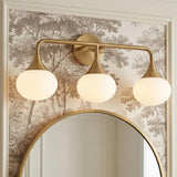A traditional bathroom featuring the Talia 3 Light Vanity, Aged Brass with white glass globes mounted above a round gold mirror on neutral tree-patterned wallpaper.