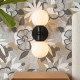 A contemporary bedroom featuring the Theo 2 Globe Wall Sconce, Matte Black with frosted glass globes mounted on floral wallpaper beside a cream textured headboard.
