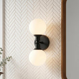 A modern bathroom with a matte black Theo 2 Globe Wall Sconce featuring frosted glass globes mounted on a white herringbone tile wall beside a wood-framed mirror.