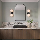 A modern bathroom with two Theo 2 Globe Wall Sconces in matte black flanking a black arched mirror above a marble countertop and fluted wood vanity.